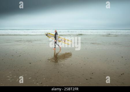 Ein einsamer Surfer mit seinem Surfbrett, der an einem kühlen nebeligen Tag in Newquay in Cornwall in t an der Küste entlang wandert und auf das Meer blickt Stockfoto