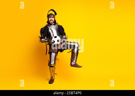 Mittelalterlicher Ritter in voller Rüstung hält Fußball auf lebendigem gelbem Hintergrund Stockfoto