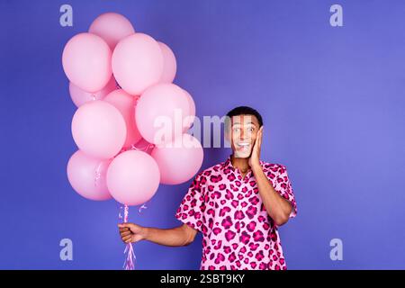 Fröhlicher Mann mit rosa Ballons vor leuchtendem lila Hintergrund, der Überraschung und Glück ausdrückt Stockfoto