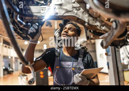 Junger Mechaniker, der in einer modernen Autowerkstatt mit Taschenlampe und Tablett das Fahrwerk inspiziert Stockfoto