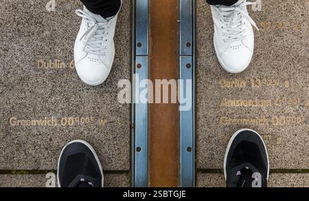 Hochwinkelaufnahme von zwei Personen, die zwischen dem Greenwich Prime Meridian stehen, markiert durch den Messingstreifen auf dem Boden im Innenhof des Royal... Stockfoto