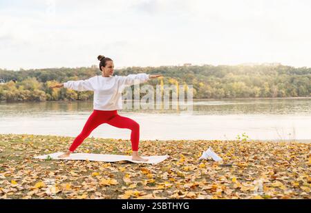 Junge Erwachsene kaukasische ethnische Frau, die im Herbst im Freien am Flussufer Yoga praktiziert. Stockfoto