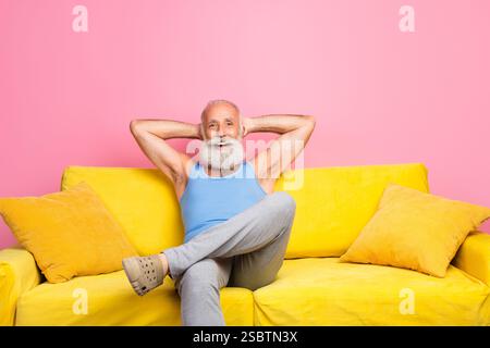 Lächelnder reifer Mann in lässiger Kleidung, der sich drinnen auf einem bunten gelben Sofa vor einer pinkfarbenen Wand entspannt Stockfoto