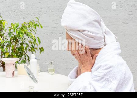 Eine ältere Frau in einem weißen Gewand und mit einem Handtuch auf dem Kopf pflegt ihre Haut vor einem Spiegel Stockfoto