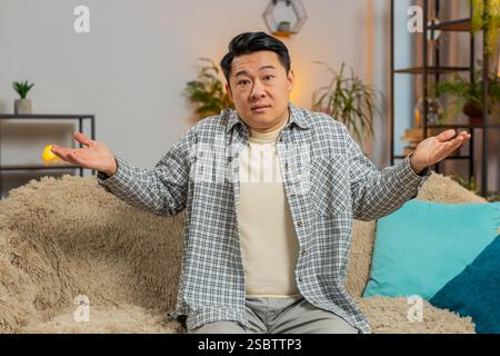 Ich weiß nicht, was ich sagen soll. Verwirrter, gutaussehender asiatischer Mann, der sich über zweideutige Fragen peinlich fühlt, Zweifel hat, keine Ahnung hat, zu Hause ahnungslos und unsicher ist. Der Chinese sitzt auf dem Sofa im Wohnzimmer Stockfoto