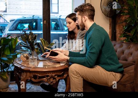 Zwei junge Profis arbeiten in einem gemütlichen Café zusammen und arbeiten an einem Laptop und Tablet. Sie konzentrieren sich auf ihre Diskussion, repräsentieren Teamwork, REM Stockfoto