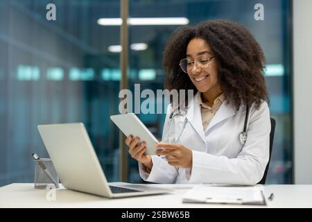 Erfahrene Ärztin, die im Büro mit Tablet-Computer und Laptop arbeitet. Die Ärztin sitzt am Schreibtisch in der Klinik und lächelt zufrieden. Stockfoto