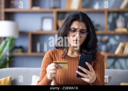 Kreditkarten- und Rechnungsprobleme. Foto einer fröhlichen jungen verwirrten Indianerin, die zu Hause ist und schockiert auf den Bildschirm des Mobiltelefons schaut. Stockfoto