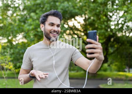 Mann, der Ohrhörer mit Smartphone im Park trägt. Aktive Konversation und Fitness im Freien. Freizeit, Technologie, Kommunikation, Natur. Stockfoto