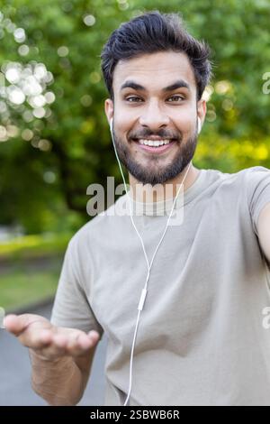 Ein Nahaufnahme-Video eines jungen arabischen Mannes mit Kopfhörern, der in einem Park steht und während eines Videoanrufs spricht, während er mit den Händen Gesten trägt. Stockfoto