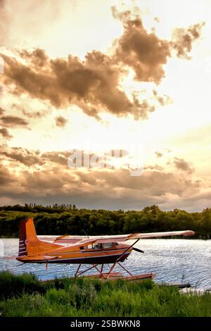 Ein kleines Flugzeug sitzt auf dem Wasser. Das Flugzeug ist orange und hat einen Propeller. Der Himmel ist bewölkt und die Sonne untergeht Stockfoto