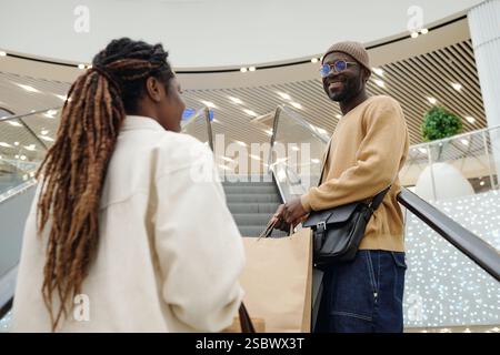 Glücklicher junger afroamerikanischer Mann in Casualwear, der seine Freundin ansieht, während beide auf der Rolltreppe im Handelszentrum stehen Stockfoto