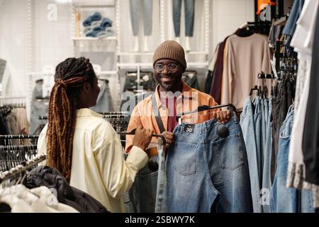 Glücklicher junger Mann, der einen Kleiderbügel mit blauer Jeans hält und seine Freundin ansieht, die ihm dabei hilft, neue Kleidung zu wählen Stockfoto