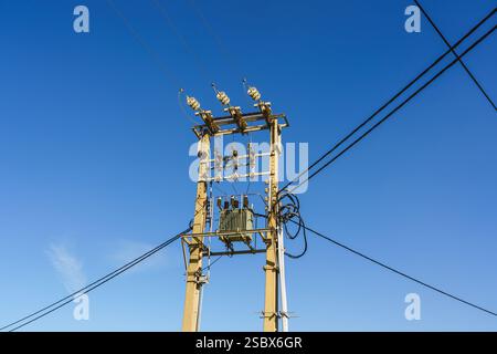 Hochspannungsleitungen und Transformator an einem Versorgungsmast unter hellblauem Himmel. Stockfoto