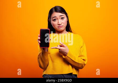 Porträt einer jungen Dame, die in die Kamera blickt und auf ein mobiles Gerät mit leerem Bildschirm zeigt. Weibliche Person, die vor einem isolierten Hintergrund steht und mit dem Zeigefinger in Richtung ihres Smartphones bewegt. Stockfoto