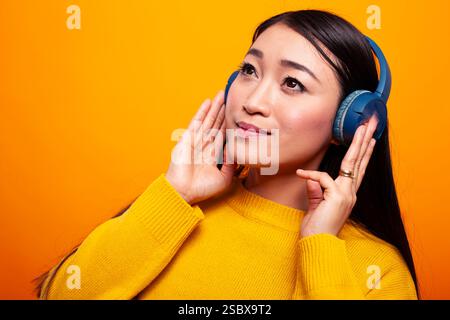 Asiatische Frau, die Musik hört, mit Kopfhörern an, genießt ihr Lieblingslied vor gelbem Hintergrund. Junge weibliche Influencerin, die ihr schnurloses Headset verwendet und wunderschöne Audiogeräusche hört. Stockfoto