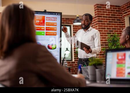 Geschäftsmann mit Zwischenablage in der Hand, mit digitalem Monitor, um Analysezusammenfassung in Besprechungen zu erläutern. afroamerikanischer Unternehmer, der mit seinem Team Projektpräsentationen und Marketingstrategien plant. Stockfoto