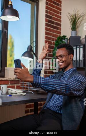 Schwarzer männlicher Profi, der Ohrstöpsel trägt, begrüßt einen Kollegen von einem entfernten Standort während eines Videoanrufs. Der afroamerikanische Manager winkt seinem Mobiltelefon zu und führt virtuelle Gespräche mit seinem Kollegen. Stockfoto