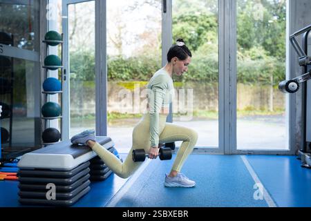 Die junge Sportlerin macht bulgarische Split Squats mit Kurzhanteln in einem Fitnessstudio, trainiert Beine und Gesäß mit einem Stepper für Balance und Unterstützung Stockfoto