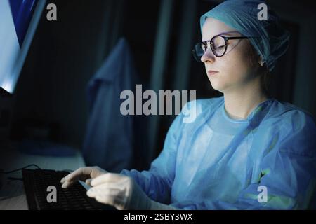Ein medizinischer Mitarbeiter in blauem Kleid, der an einem Computer bei schwacher Beleuchtung arbeitet Stockfoto