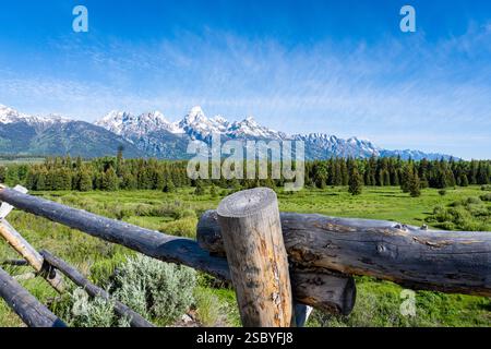 Ein atemberaubender Blick auf die Grand Tetons mit einem rustikalen Zaun, üppigem Grün, blauem Himmel und schneebedeckten Gipfeln. Stockfoto