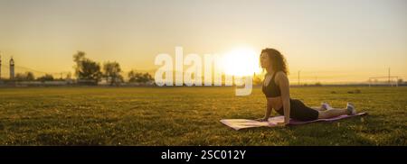 Eine Frau, die Kobra Yoga macht, posiert auf einer Matte auf einem weiten, ruhigen Feld während des Sonnenaufgangs Stockfoto