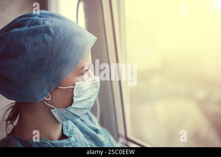 Ein medizinischer Mitarbeiter in Schutt und Maske blickt bei Sonnenuntergang aus dem Fenster und wirkt kontemplativ Stockfoto