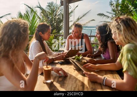 Verschiedene Frauen speisen im tropischen Restaurant am Meer. Weibliche Freunde lachen in der Nähe von Palmen. Geselliges Essen, Urlaubsstimmung, Sitzplätze im Freien Stockfoto