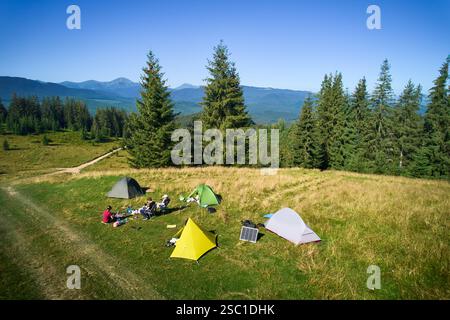 Campingplatz aus der Vogelperspektive in den Bergen, mit mehreren bunten Zelten auf einer grasbewachsenen Rodung. Camper entspannen unter klarem blauen Himmel, umgeben von hohen immergrünen und Panoramablick auf entfernte Gipfel. Stockfoto