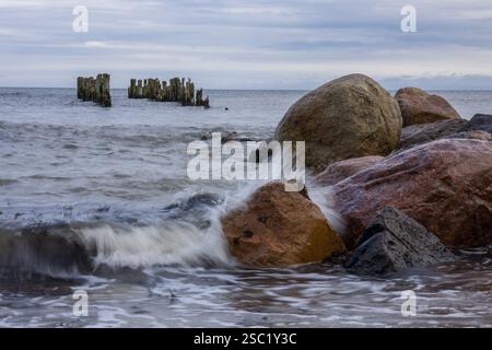 Dramatische Meereslandschaft mit Wellen, die gegen Felsen krachen und verwitterten Holzpfosten im Hintergrund. Kraftvolle und ungezähmte Naturszene. Perfekt für Natur Stockfoto