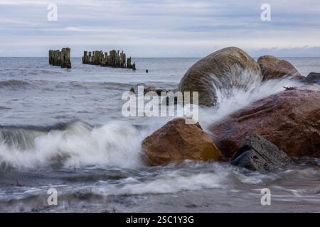 Dramatische Meereslandschaft mit Wellen, die gegen Felsen krachen und verwitterten Holzpfosten im Hintergrund. Kraftvolle und ungezähmte Naturszene. Perfekt für Natur Stockfoto