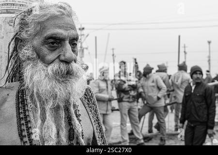 14. Januar 2025, Prayagraj, uttar Pradesh, Indien. Ein langbärtiger Sadhu blickt nach vorne, während die Polizei im Hintergrund steht und den Kontrast symbolisiert Stockfoto