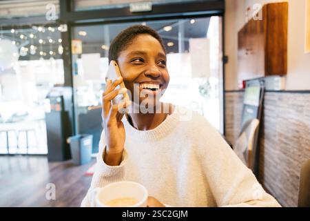 Eine junge Frau genießt eine Kaffeepause und plaudert fröhlich auf ihrem Smartphone in einem gemütlichen Café. Stockfoto