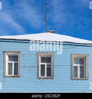 Fenster in dekorativen geschnitzten Rahmen sind in alter blauer Holzhauswand unter blauem Himmel an einem sonnigen Wintertag, quadratische architektonische Hintergrundtextur, f Stockfoto