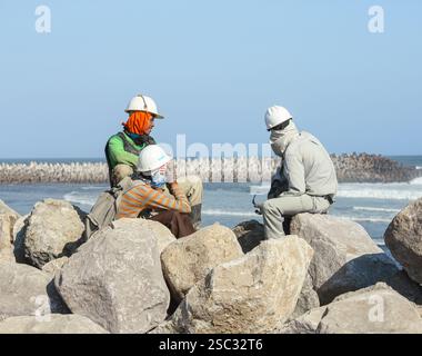 Drei Bauarbeiter in weißen Schutzhüten sitzen auf großen Felsen und genießen eine Pause am Meer mit Blick auf Tetrapoden und den klaren blauen Himmel Stockfoto