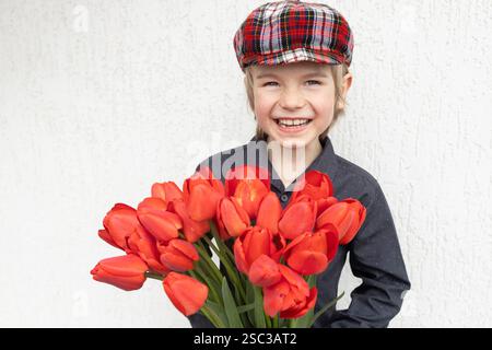 Porträt eines niedlichen Jungen in der Kappe 6, 7 Jahre alt, lächelnd fröhlich, in Händen viele rote Tulpen auf hellem Hintergrund. Muttertag. Valentinstag. Ha Stockfoto