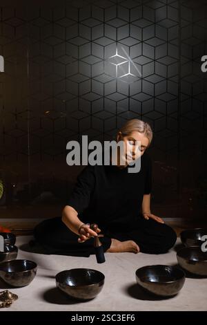 Eine Frau sitzt ruhig auf dem Boden und übt Klangheilung mit mehreren Klangschalen, die um sie herum angeordnet sind. Die Einstellung umfasst sanfte Beleuchtung und pla Stockfoto