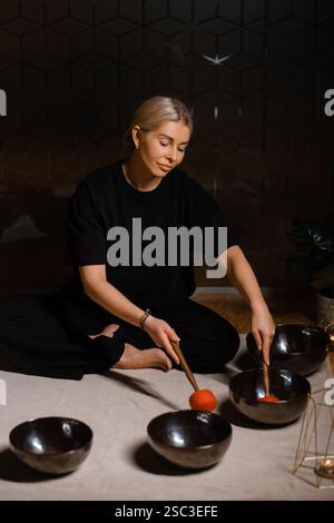 Eine Frau sitzt ruhig auf dem Boden und übt Klangheilung mit mehreren Klangschalen, die um sie herum angeordnet sind. Die Einstellung umfasst sanfte Beleuchtung und pla Stockfoto