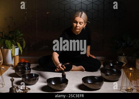 Eine Frau sitzt ruhig auf dem Boden und übt Klangheilung mit mehreren Klangschalen, die um sie herum angeordnet sind. Die Einstellung umfasst sanfte Beleuchtung und pla Stockfoto