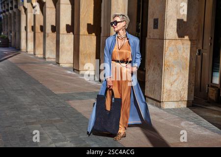 Stilvolle Seniorin stehend City Street in modischen Kleidern, trägt langes beigefarbenes Seidenkleid, blauen Cape Coat, trägt Maxi Tote schwarze Handtasche, Sonne Stockfoto