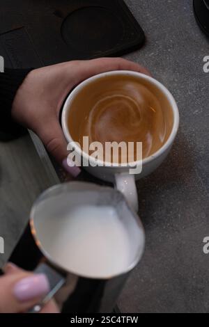 Draufsicht eines weiblichen Barista, der aufgeschäumte Milch aus einem Metallkrug in eine weiße Tasse gießt und so Latte Art kreiert. Kaffeezubereitungsprozess. Stockfoto