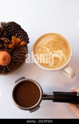 Blick von oben auf einen Portafilter mit gemahlenem Kaffee und einer Tasse Latte mit Kunst. Die Hand des Barista hält die Ausrüstung auf einem weißen Hintergrund. Stockfoto