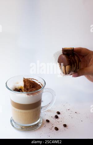 Hand hält ein kleines Schokoladendessert neben einem Glas Lagenkaffee mit Schaum und Kakao. Sweet Break und Energy Boost Konzept. Stockfoto