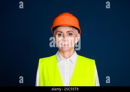 Selbstbewusste reife Frau in Baukleidung, trägt einen orangefarbenen Schutzhelm und eine Neonweste vor einem dunkelblauen Hintergrund Stockfoto