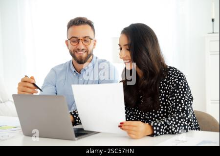 Zwei erfolgreiche positive Geschäftskollegen, die an einem Schreibtisch in einem modernen Büro mit Dokumenten arbeiten, beide lächeln, mit einem Laptop und Diagrammen auf dem Schreibtisch, die Ideen austauschen, entwickeln einen Geschäftsplan Stockfoto