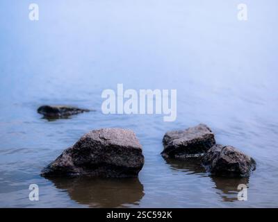 Minimalistische Zusammensetzung von Felsen, die aus ruhigem Wasser auftauchen, schaffen eine friedliche und ruhige Seeszene mit einem weichen blauen Farbverlauf. Stockfoto