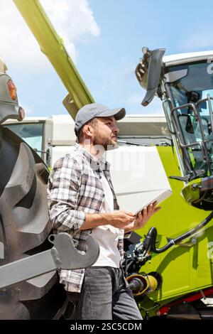 Händler oder Lieferant von Landmaschinen. Kaukasischer Mann in kariertem Hemd, der neben landwirtschaftlichen Maschinen steht und ein digitales Tablet benutzt. Stockfoto