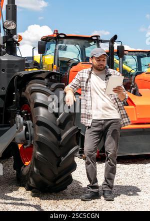 Händler oder Lieferant von Landmaschinen. Kaukasischer Mann in kariertem Hemd steht neben dem Traktor und benutzt ein digitales Tablet. Stockfoto