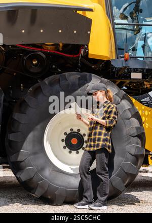 Händler oder Lieferant von Landmaschinen. Weiße Frau im karierten Hemd steht neben dem Mähdrescher und verwendet ein digitales Tablet. Stockfoto