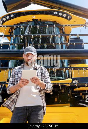 Händler oder Lieferant von Landmaschinen. Kaukasischer Mann in kariertem Hemd steht neben Mähdrescher und verwendet ein digitales Tablet. Stockfoto